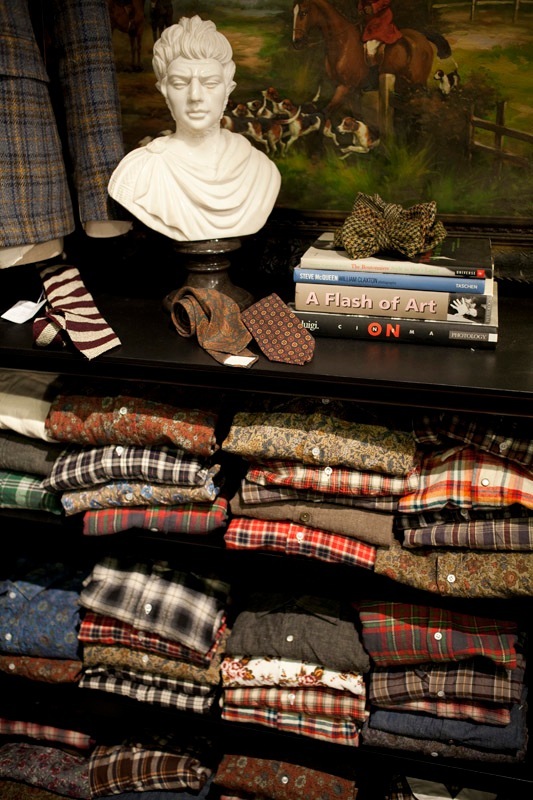 Folded Salvatore Piccolo work shirts in various plaid and checked patterns displayed on shelves with a white bust and art books.
