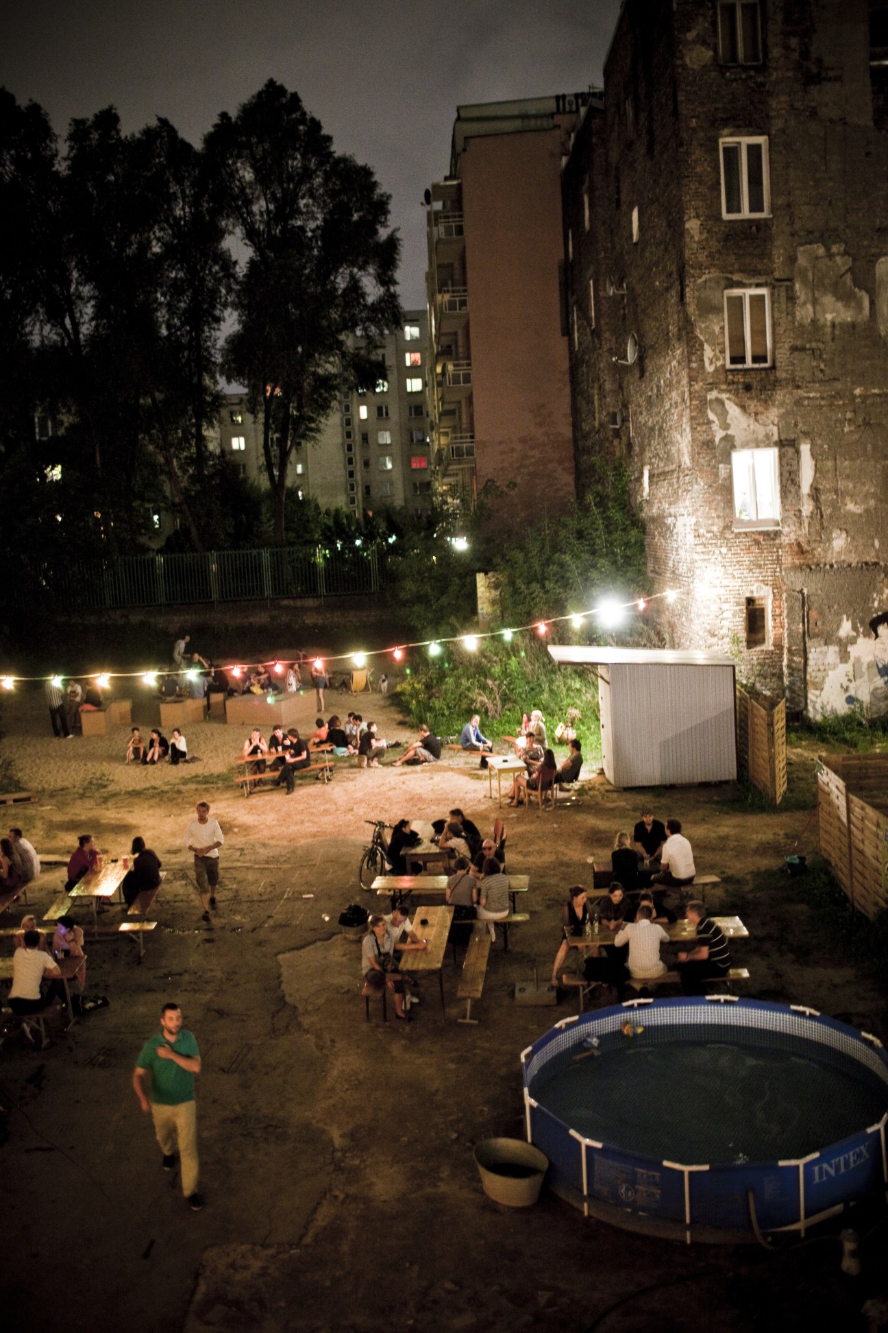Evening gathering at 5.10.15 night spot with people socializing around tables in a courtyard lit by string lights and a swimming pool in foreground.