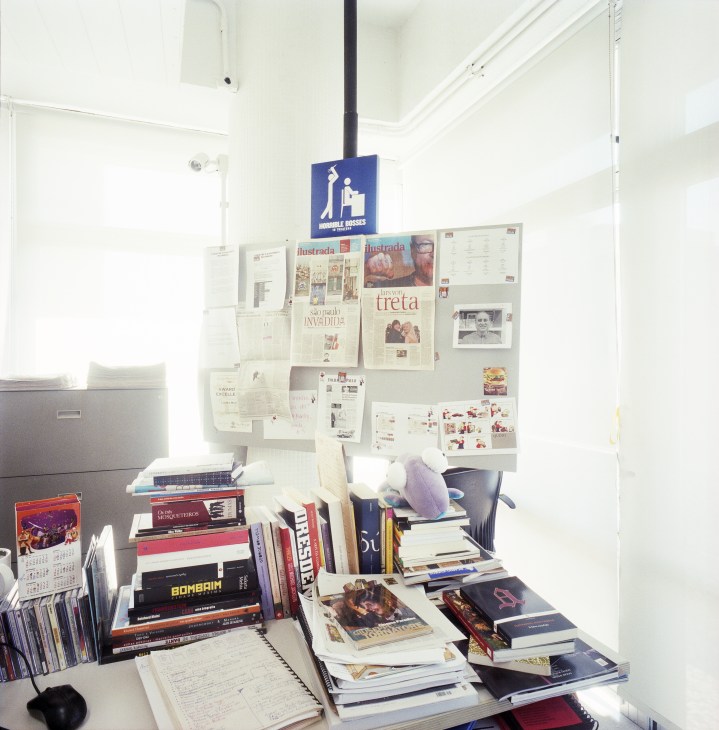A cluttered desk overflowing with books, magazines, and newspaper clippings pinned to a wall board above.