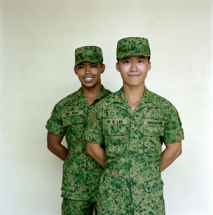 Two Singapore servicemen in green digital camouflage uniforms and caps stand side by side against a plain background.