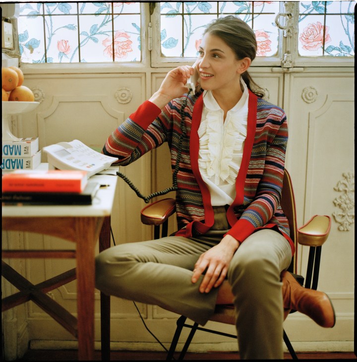 Woman sitting in an armchair wearing a John Smedley cardigan, J.Crew shirt, Loro Piana trousers, and Camper shoes, talking on a vintage telephone.