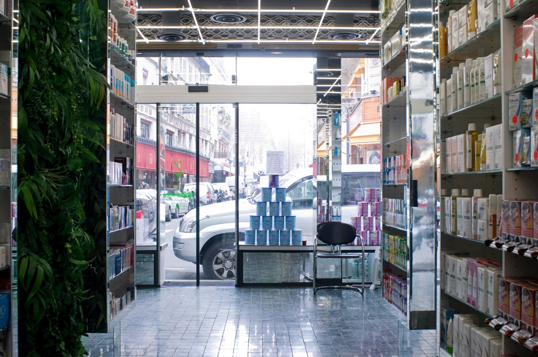 Interior of Mapharmacie pharmacy in Paris with mirrored shelves, minimal product displays, and a plant-covered stockroom doorway visible through large storefront windows.