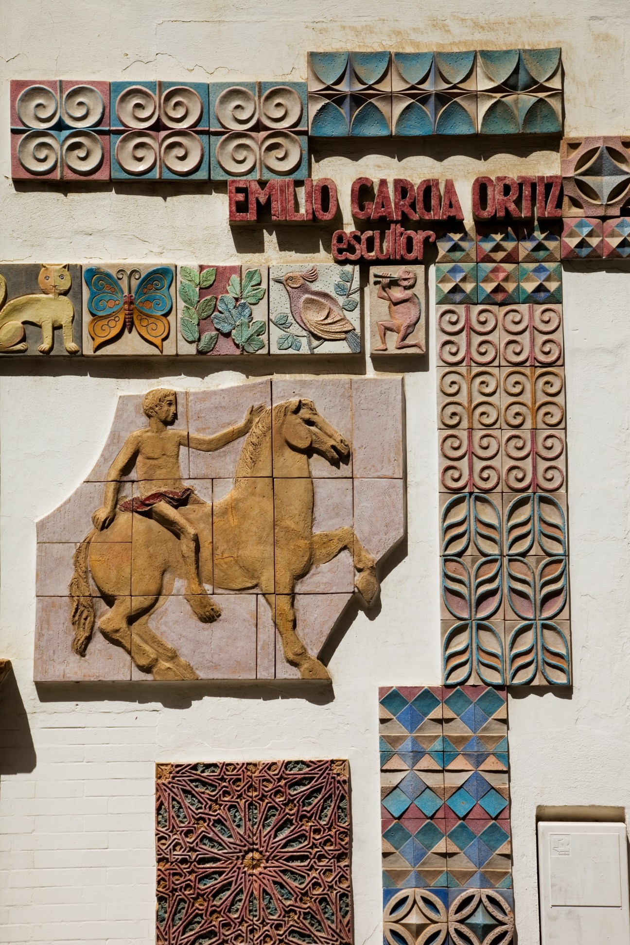 Decorative ceramic tiles and sculptural elements on a white wall, featuring traditional Spanish pottery designs with geometric patterns and figurative reliefs.