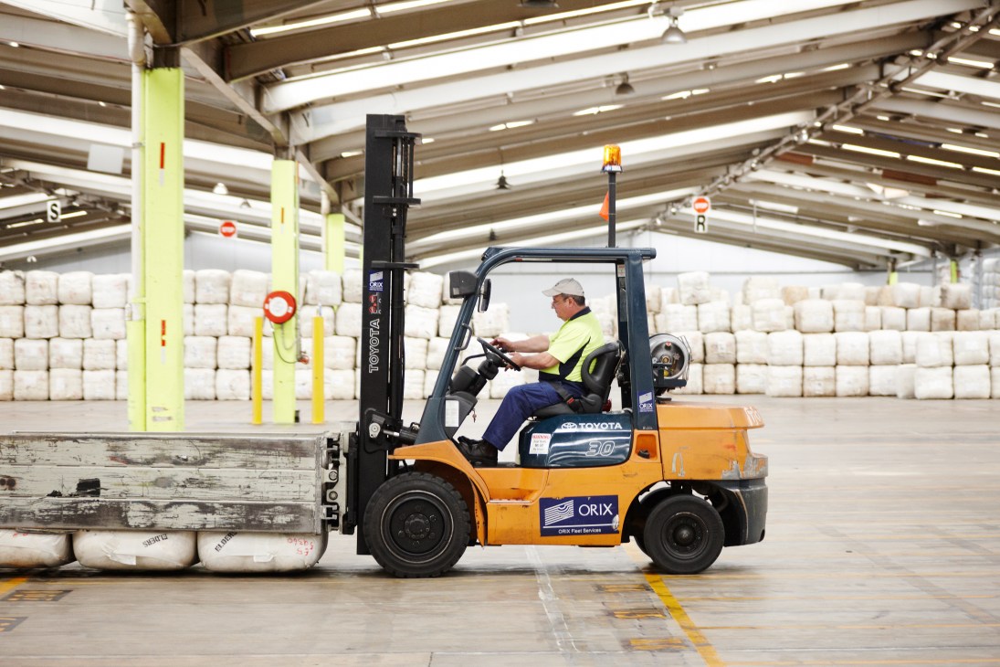 Forklift operator in yellow safety vest and hard hat operating an orange Toyota forklift to move compressed wool bales in a large warehouse facility.