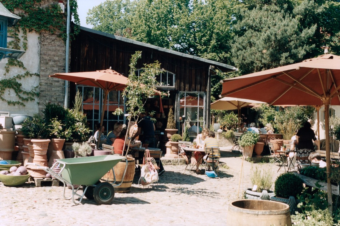 Outdoor courtyard of Mutter Fourage deli-café with terracotta umbrellas, potted plants, and customers dining.