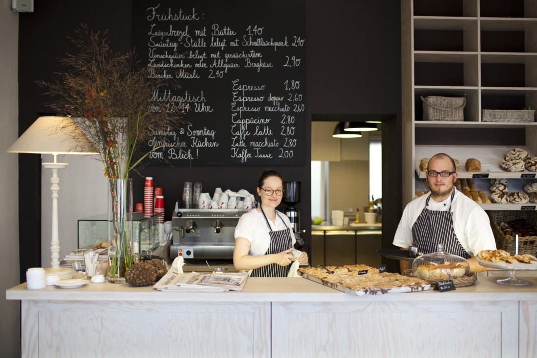 Two staff members standing behind the counter at Alpenstück bakery in Berlin's NoTo neighbourhood, with a chalkboard menu and baked goods displayed.