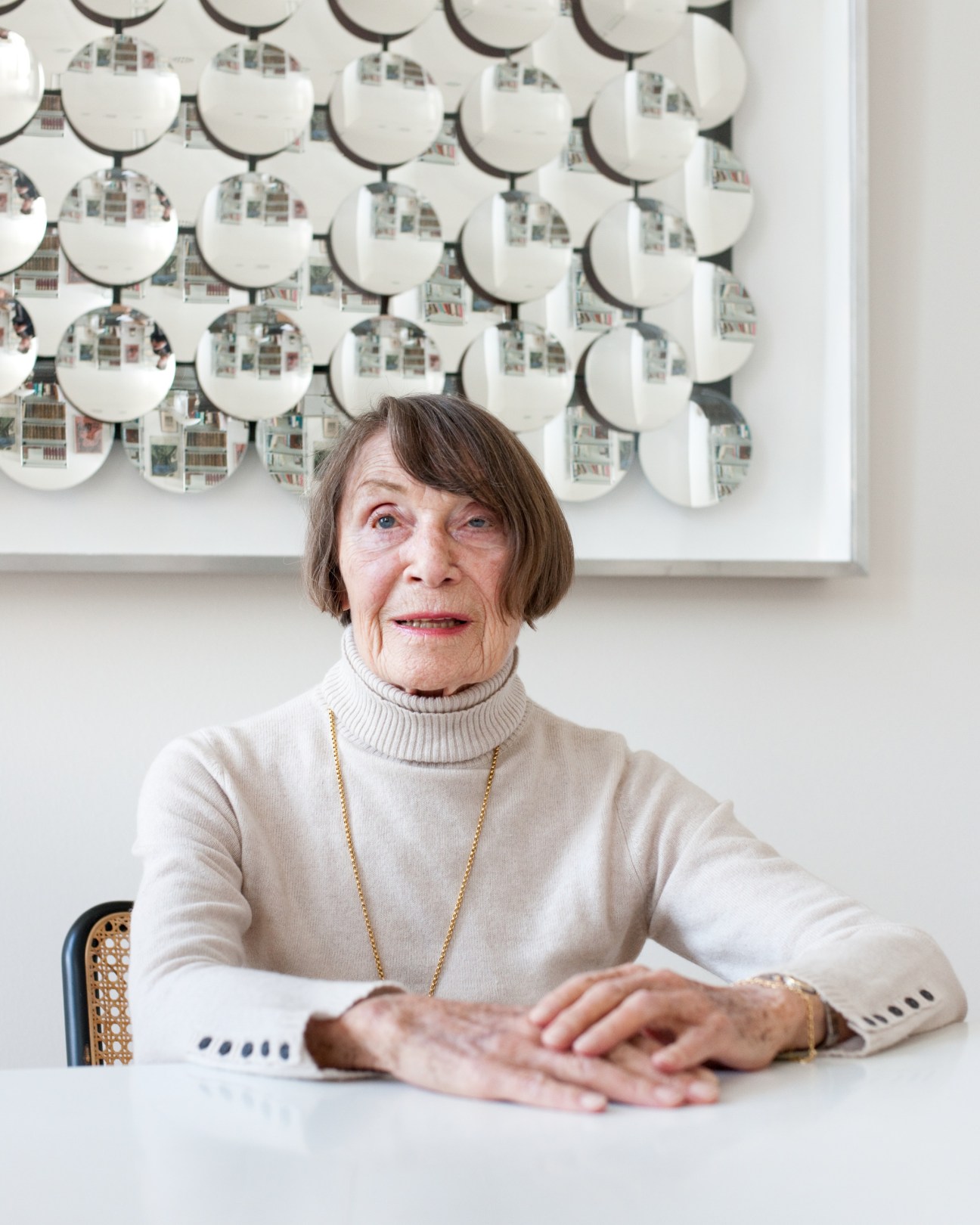 Anneliese Friedmann sits at a table wearing a cream turtleneck and gold necklace, with circular artwork displayed on the wall behind her.