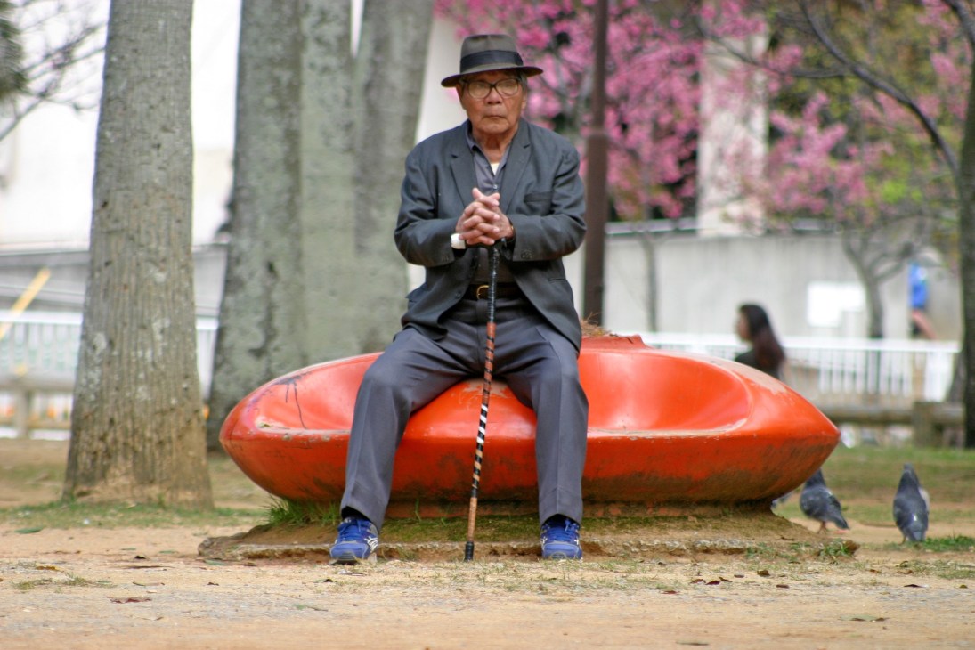 An elderly man sits on a red sculptural seat in a Japanese public space, holding a walking stick.