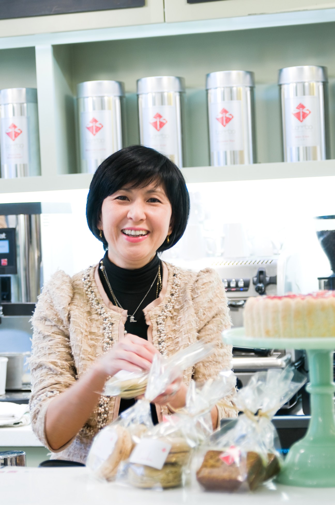 Woman in beige tweed jacket smiling at Tombo Japanese deli counter with packaged products.