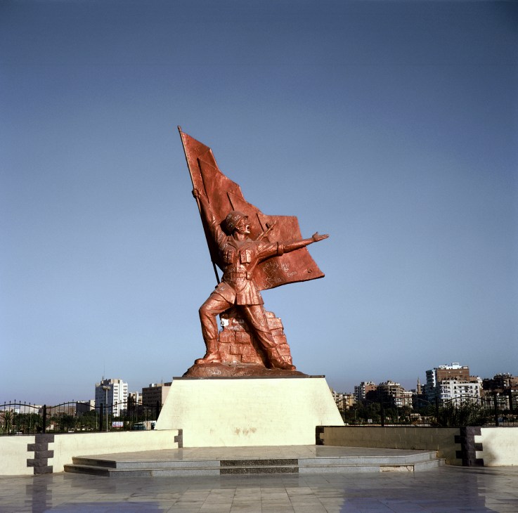 A large bronze statue of a soldier with a flag on a white stone pedestal in Suez City.