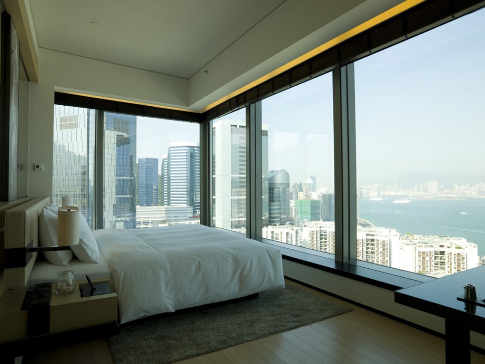 Modern hotel bedroom with floor-to-ceiling windows overlooking Hong Kong harbour and skyline.