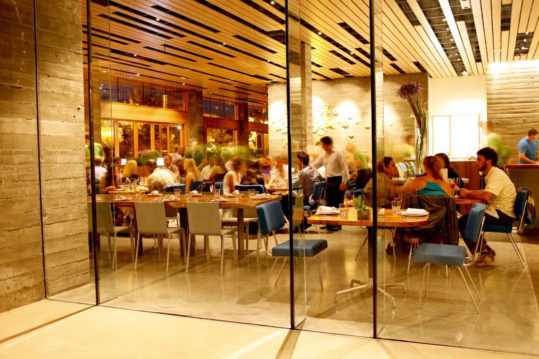 Diners seated at tables inside H2 Hotel restaurant with floor-to-ceiling glass windows and warm wooden ceiling.