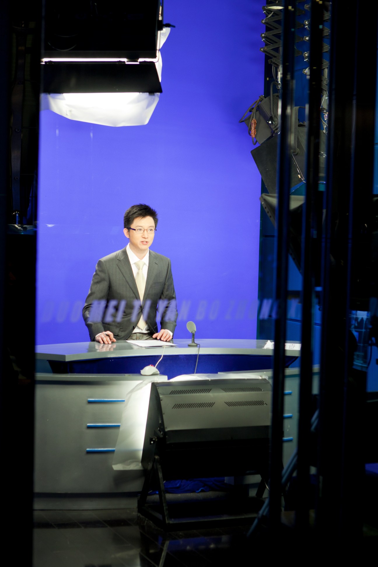 News anchor Zhang Daxin sits at a desk in a television studio with a bright blue backdrop and professional lighting.