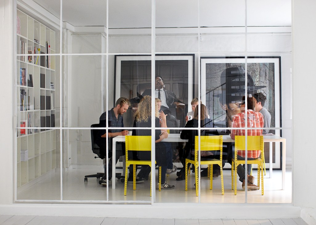 Staff members working at tables with yellow chairs in a bright, minimalist office space with glass walls and shelving.