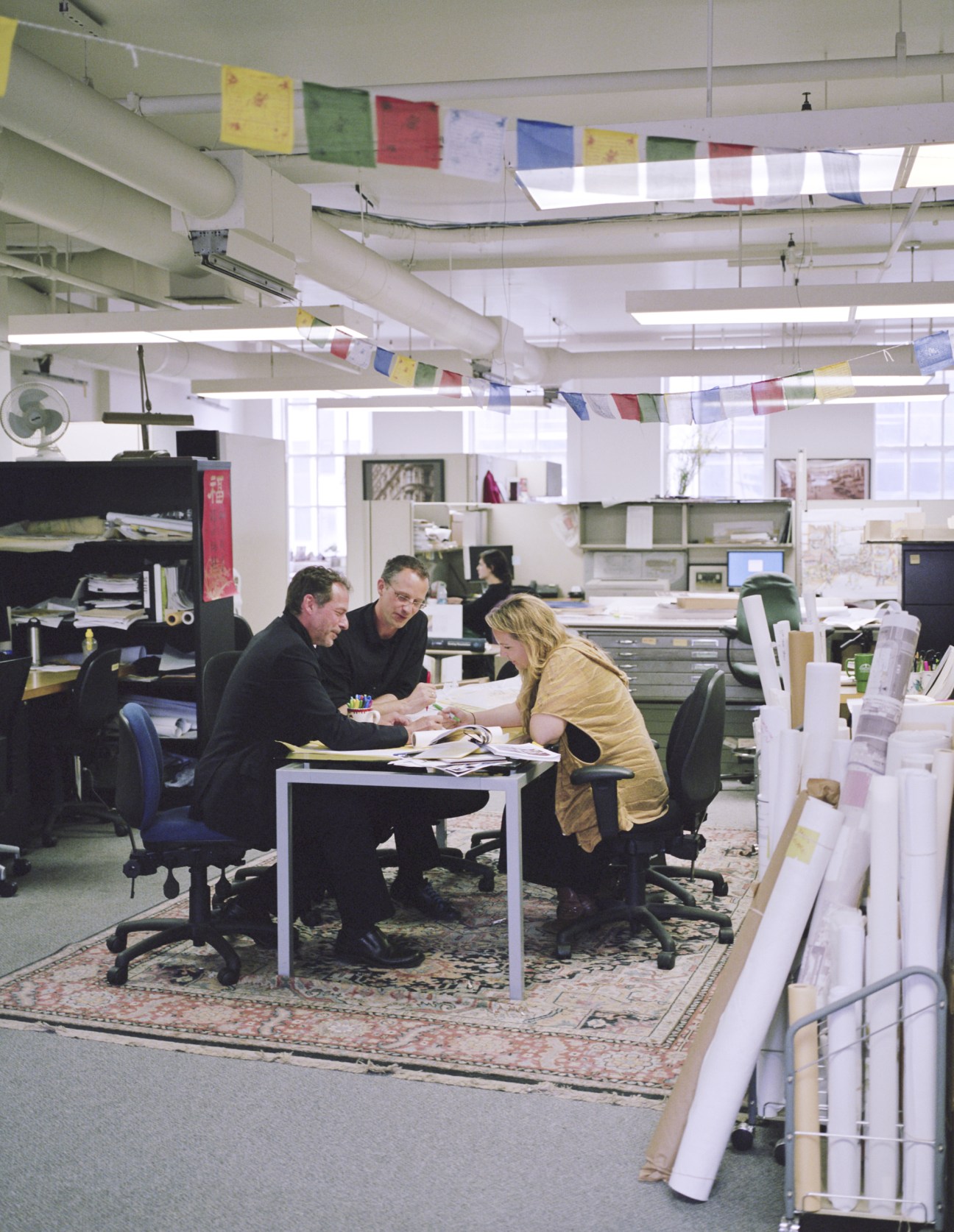 Three urban planners discuss designs at a table in a bright office studio filled with sketches, maps, and colorful prayer flags.