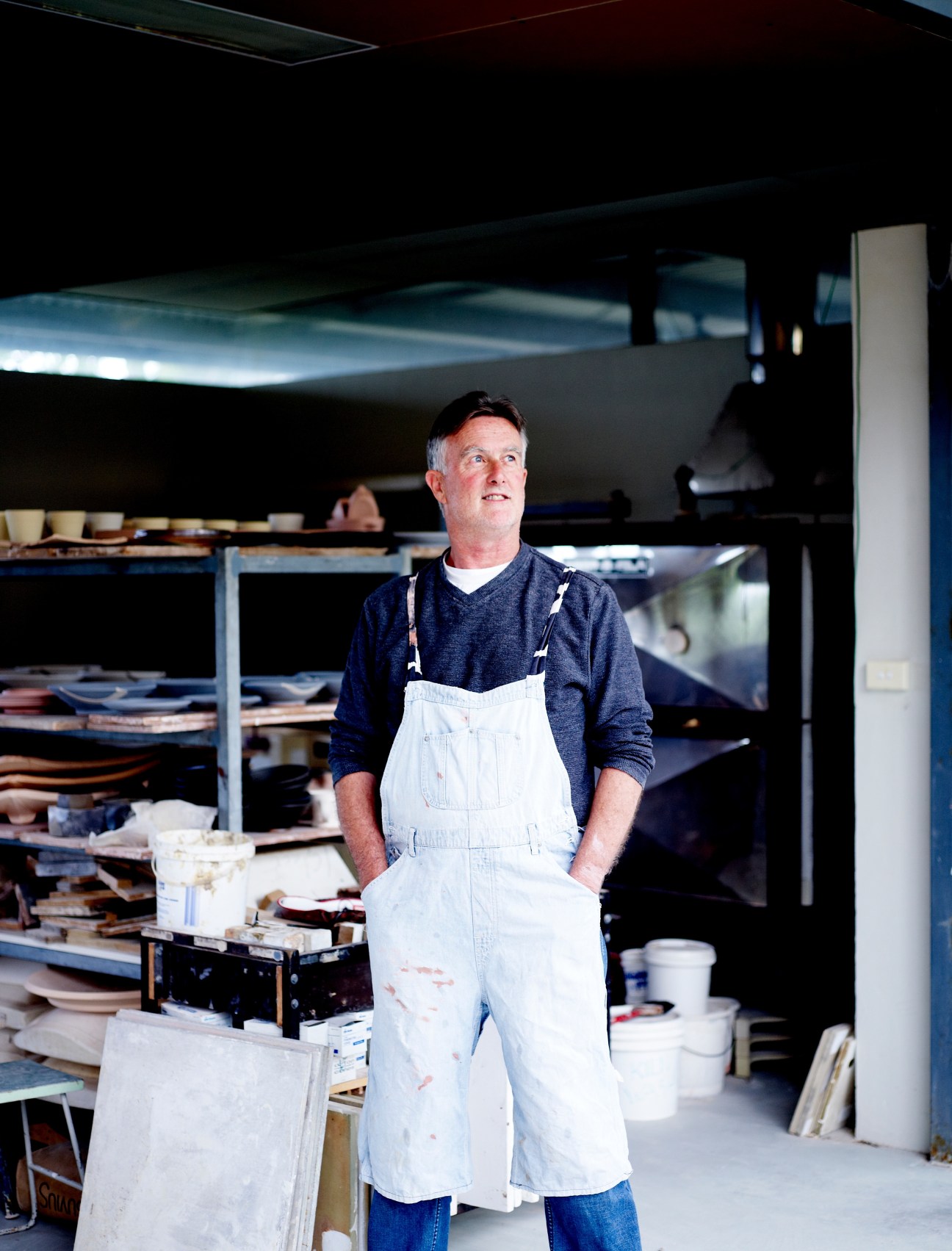 Man in white apron standing in woodworking workshop with shelves of materials.