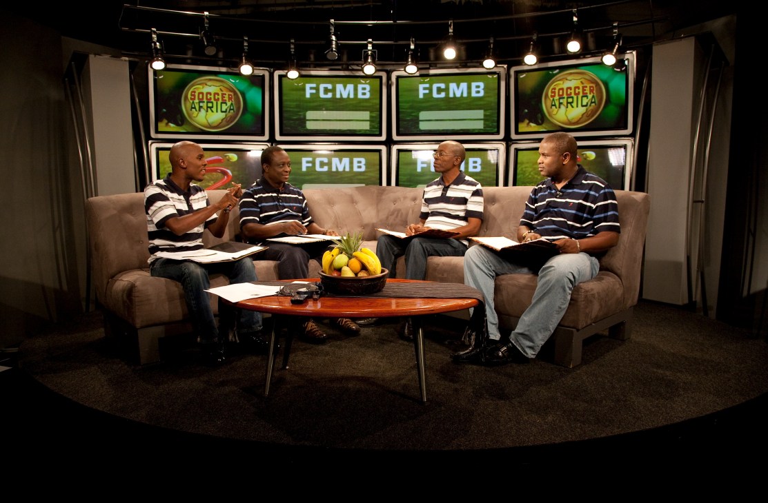 Four men in striped polo shirts sit on a beige sofa on the Soccer Africa TV set with FCMB and Soccer Africa logos on screens behind them.