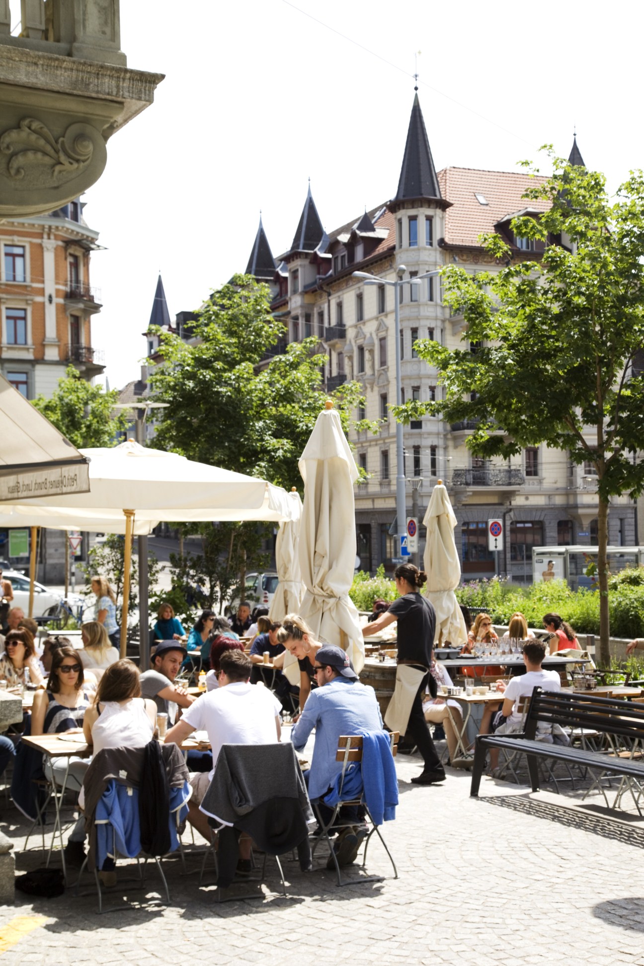Outdoor café terrace in a European city square with historic architecture and leafy trees.