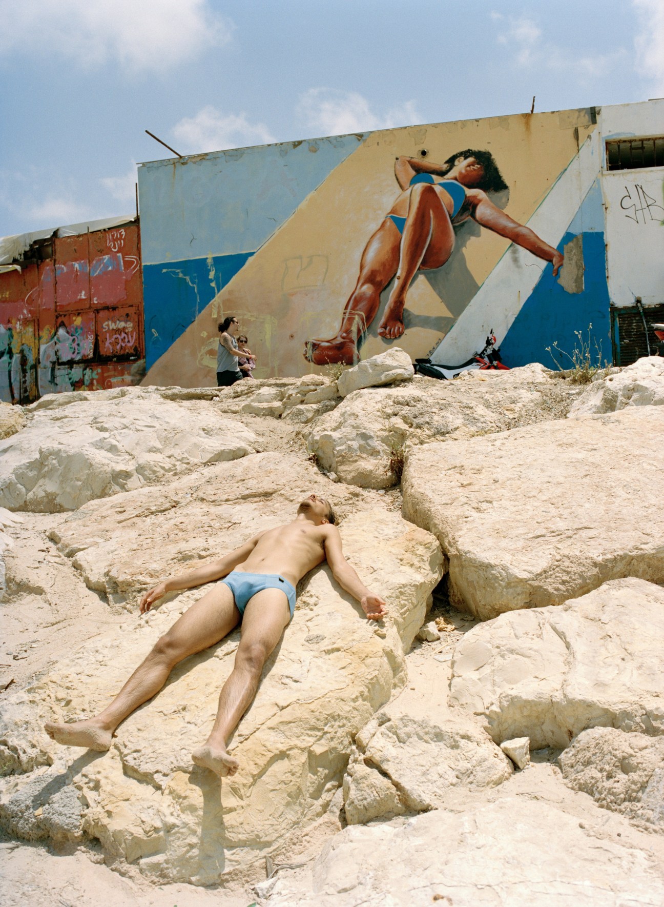 A shirtless man in blue swim shorts lies on rocky sand near a weathered blue and white building with street art depicting a woman in a blue bikini.