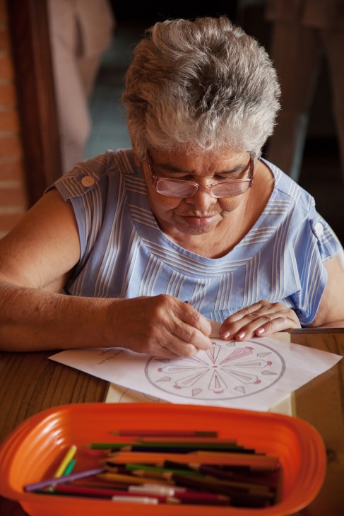 Elderly woman with white hair and glasses drawing a flower design at a table with colored pencils nearby.