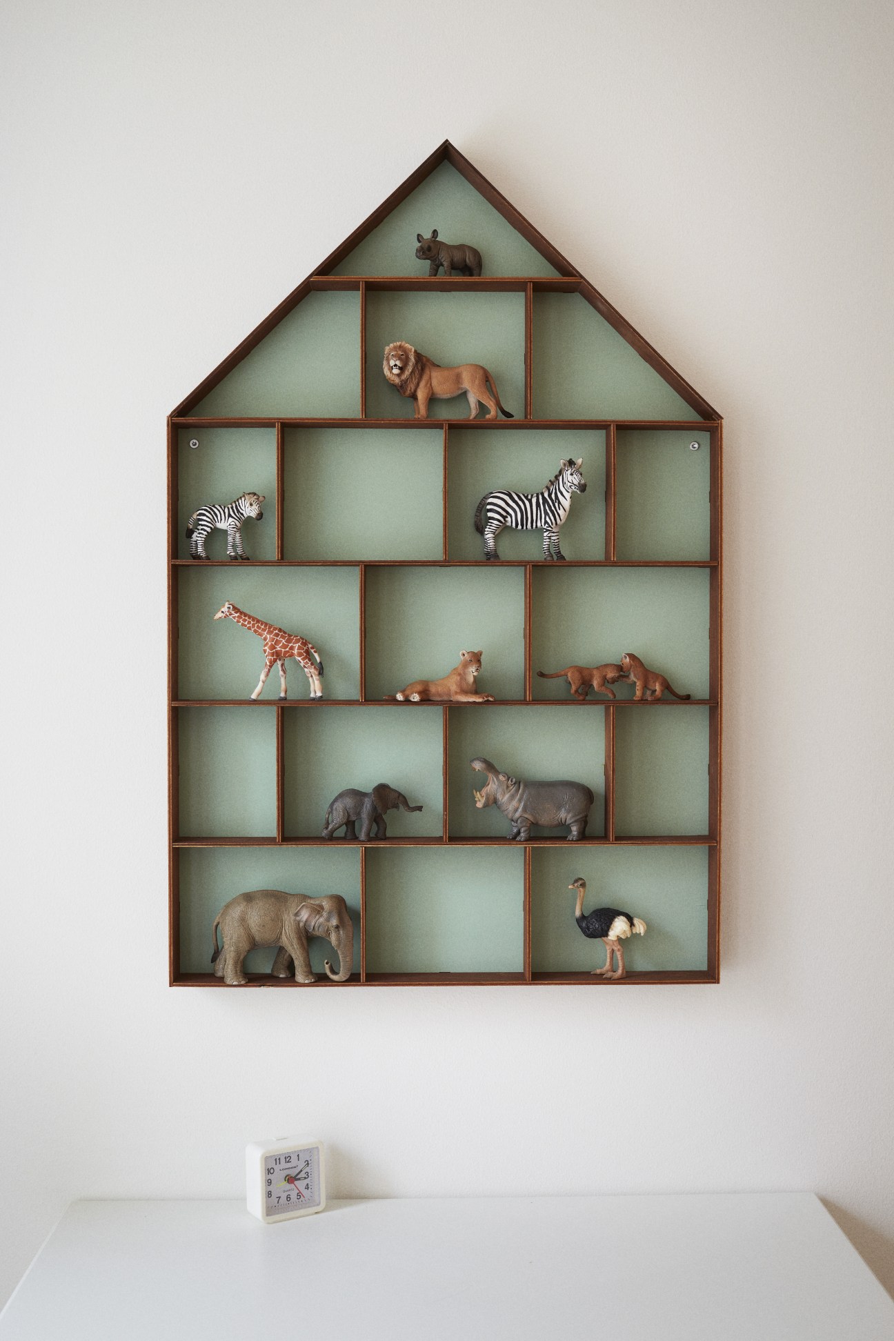House-shaped wooden shelving unit displaying animal figurines including zebras, lions, giraffes, and elephants on a sage green background.