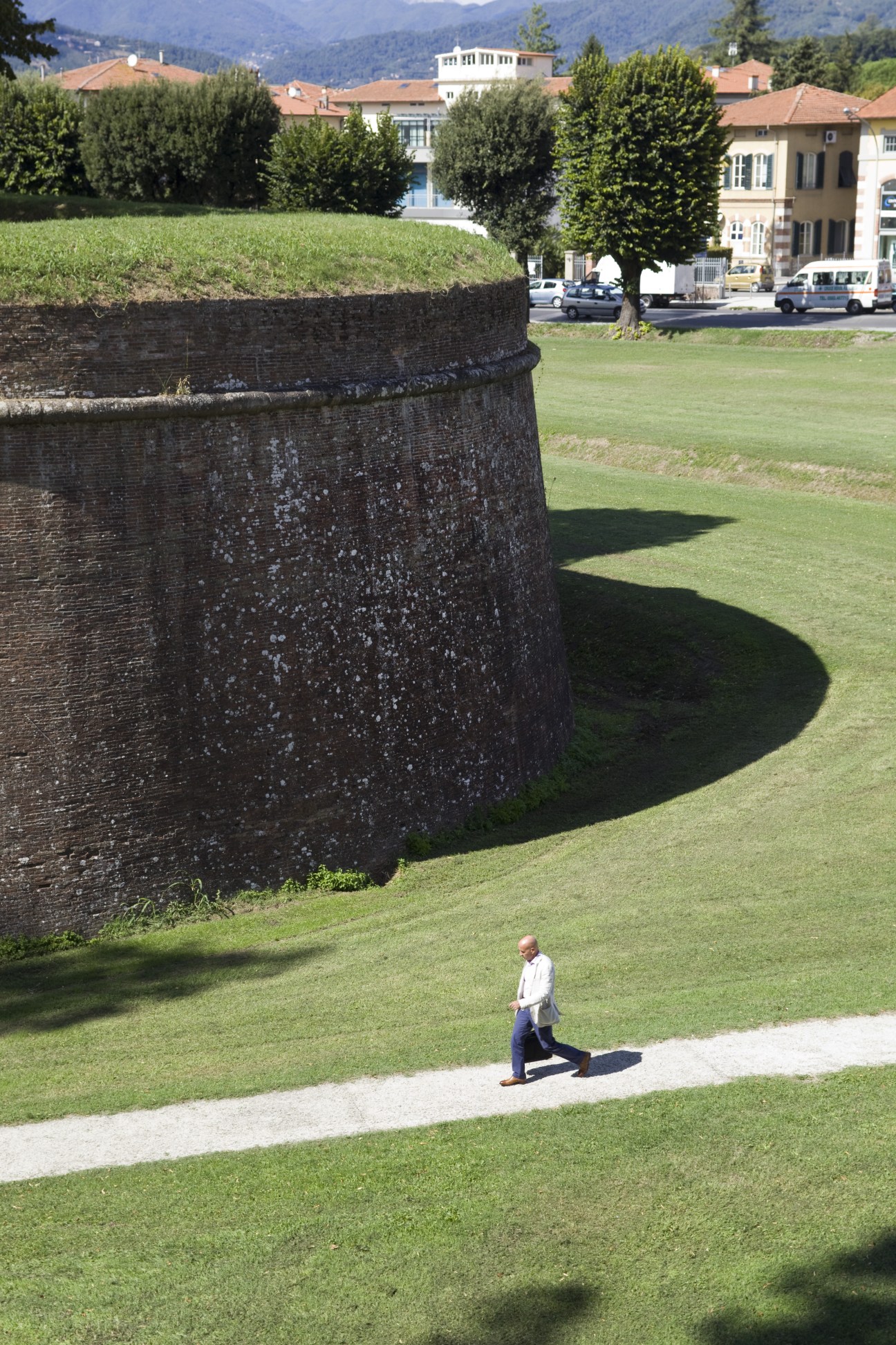 A jogger runs along a grassy path beside Lucca's imposing red-brick fortified walls with mountains visible in the background.