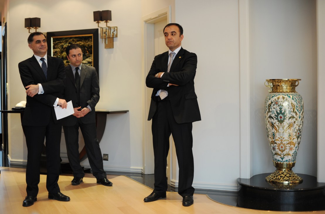 Three men in dark suits stand in an elegant foyer with classical décor and an ornamental vase.