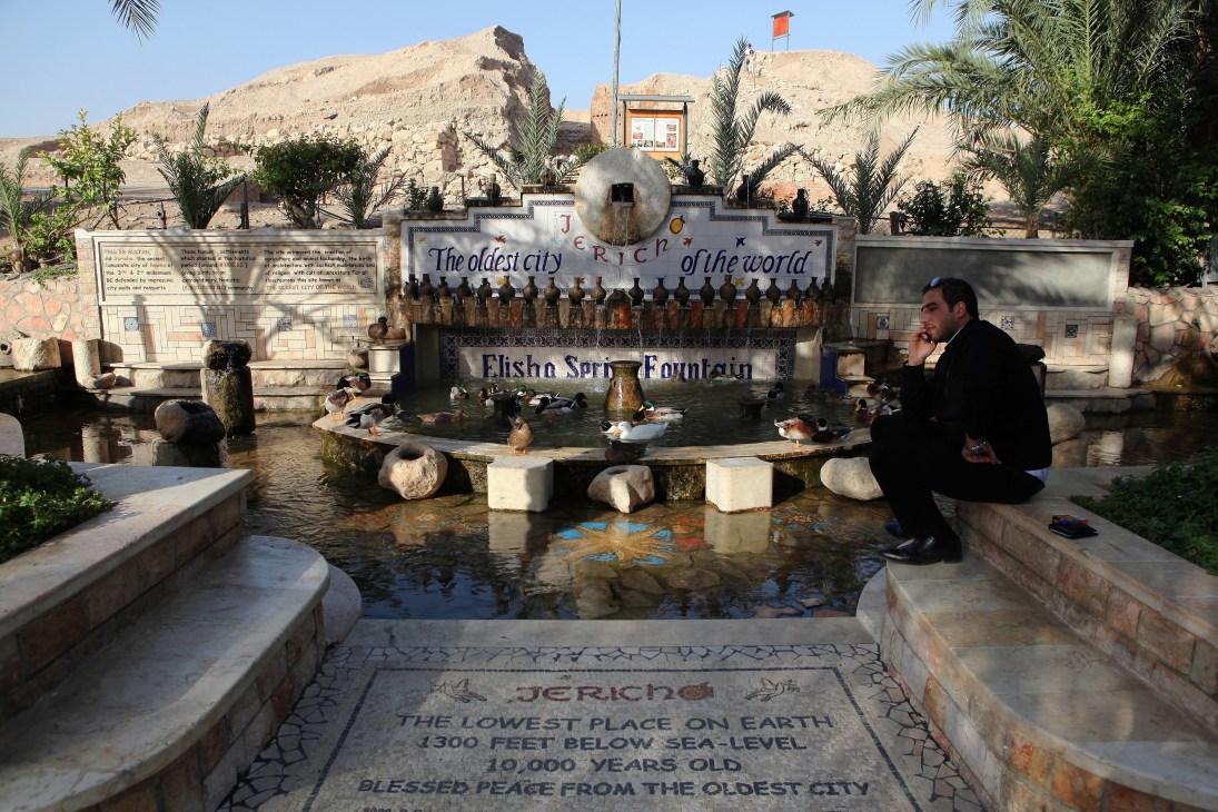 Elisha Spring fountain with signage declaring Jericho as the oldest city in the world, surrounded by palm trees and ancient ruins.