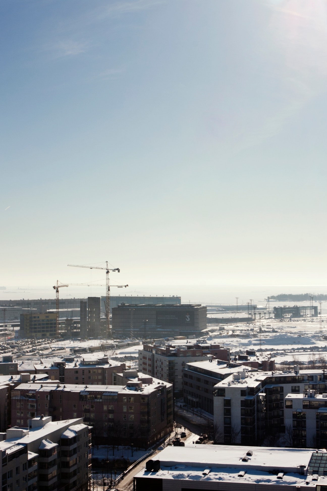 Urban construction site with cranes, low-rise residential buildings, and harbor area in background.