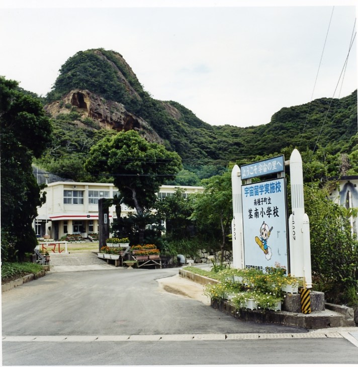 A Japanese school entrance with a mountainous backdrop and a rocket-shaped sign in the foreground.
