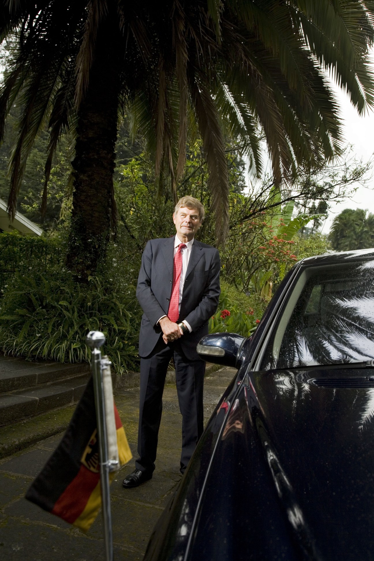 German architect Michael Maivald stands outside next to a black car with tropical plants and a German flag visible.
