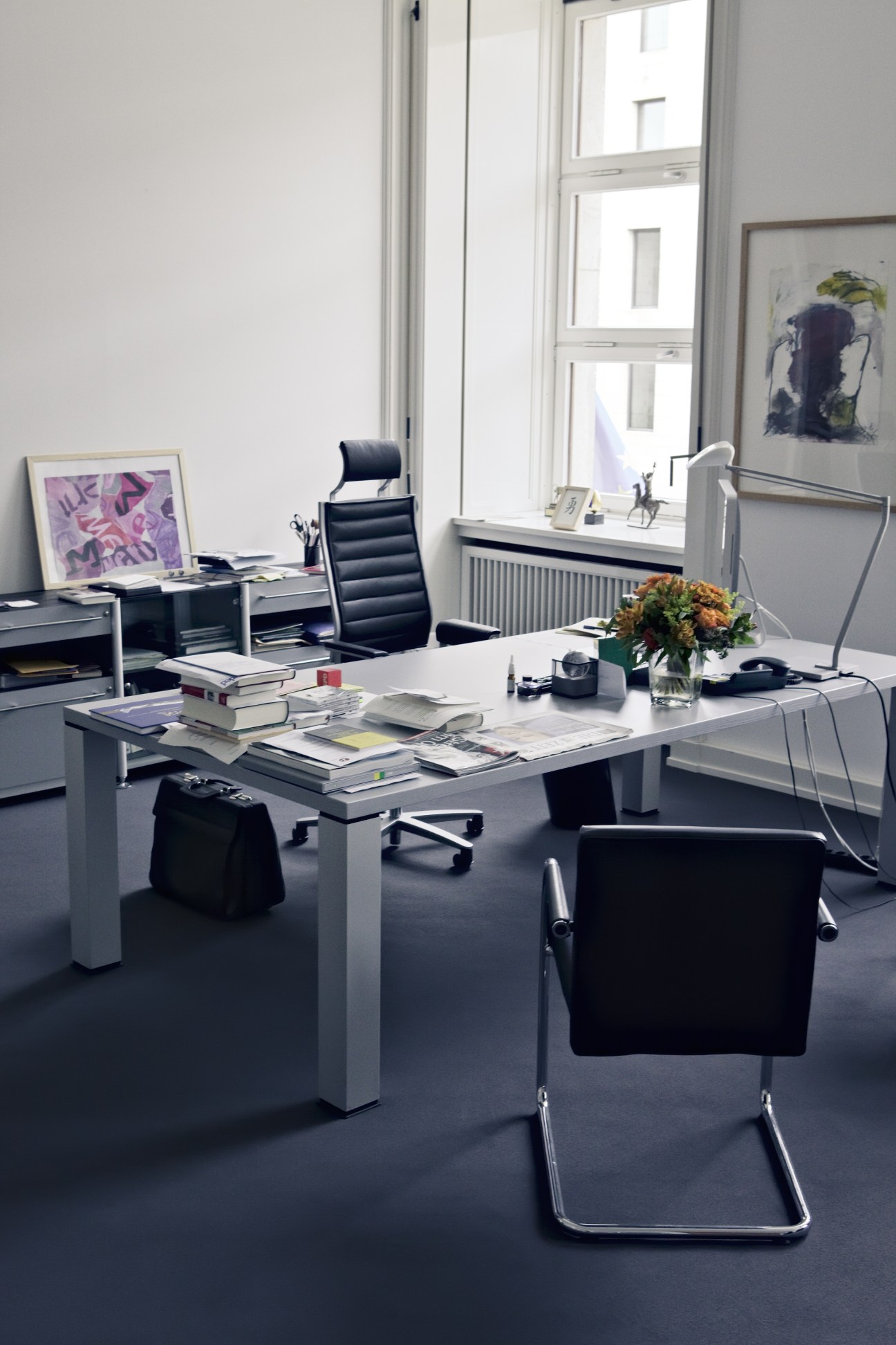 Modern ministerial office with minimalist white desk, black chairs, artwork, and windows overlooking Berlin.