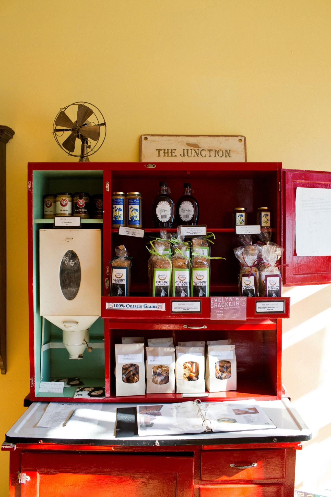 Red vintage cabinet display with artisanal chocolates, jams, and baked goods on yellow wall