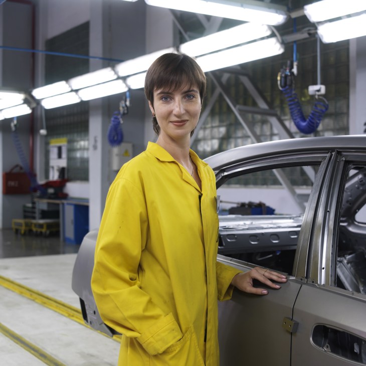 Woman in yellow work coat standing next to a car on an assembly line at Avtotor car plant.