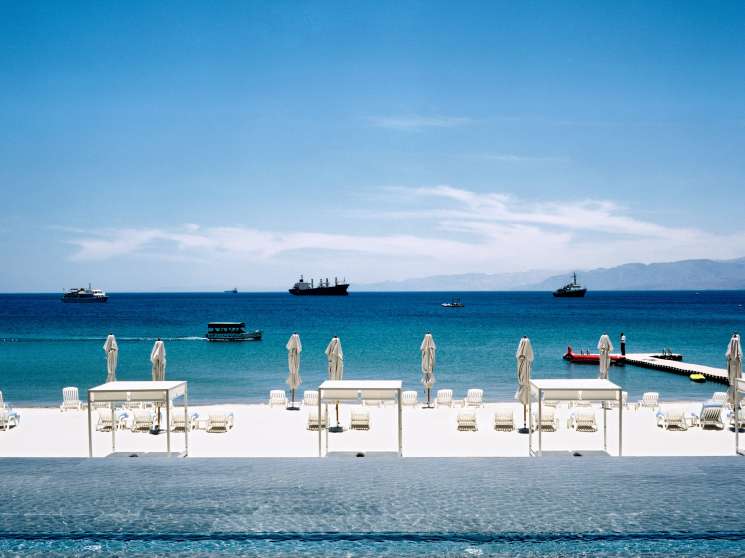White umbrellas and loungers line a pristine beach at Hotel Kempinski with turquoise waters and anchored ships visible.