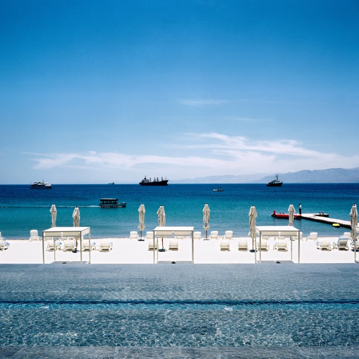 White umbrellas and loungers line a pristine beach at Hotel Kempinski with turquoise waters and anchored ships visible.