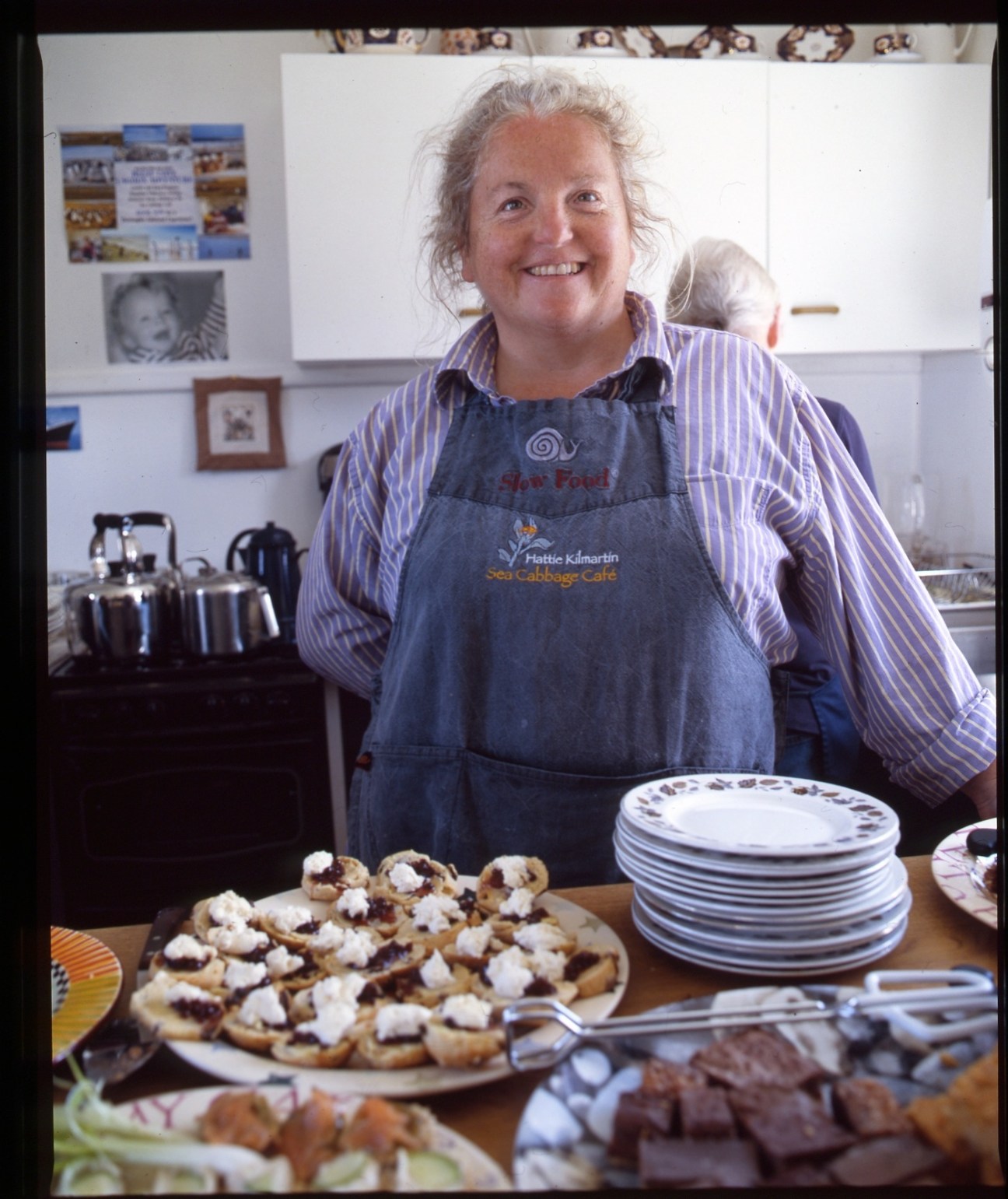Hattie Kilmartin smiles in the Sea Cabbage Café kitchen wearing an apron, with scones and refreshments displayed on the counter.