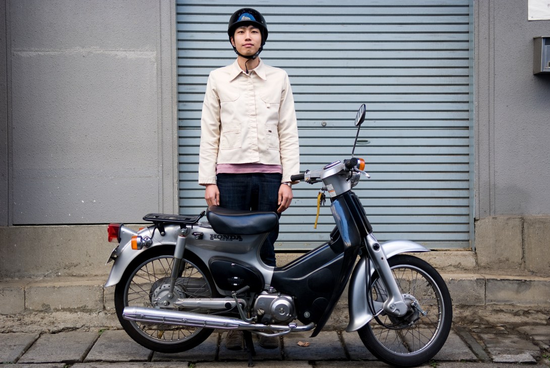Young man in white jacket and helmet standing with a silver Honda Super Cub scooter against a metal garage door.