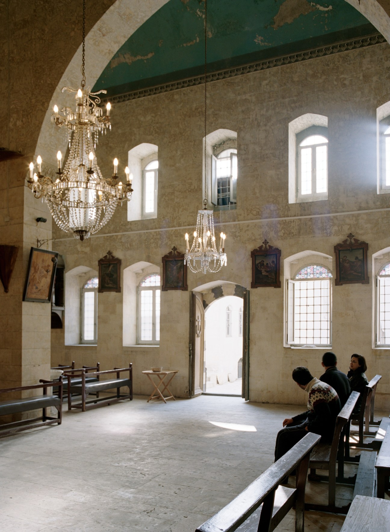 Interior of Syrian Catholic church with ornate chandeliers, arched windows, religious paintings, and visitors on wooden benches.
