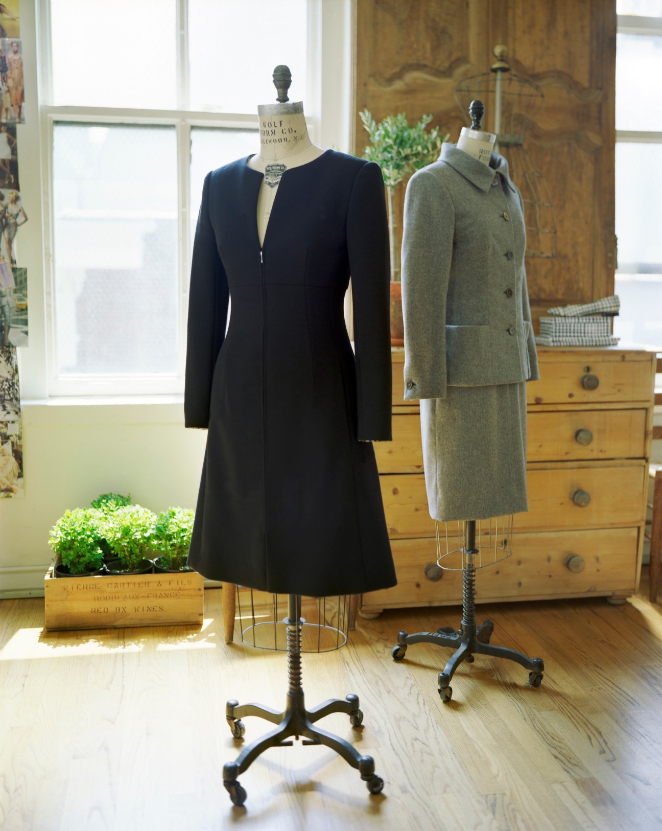 Two dress forms display Melograno womenswear: a black long-sleeved dress and a grey wool coat in a bright studio setting.