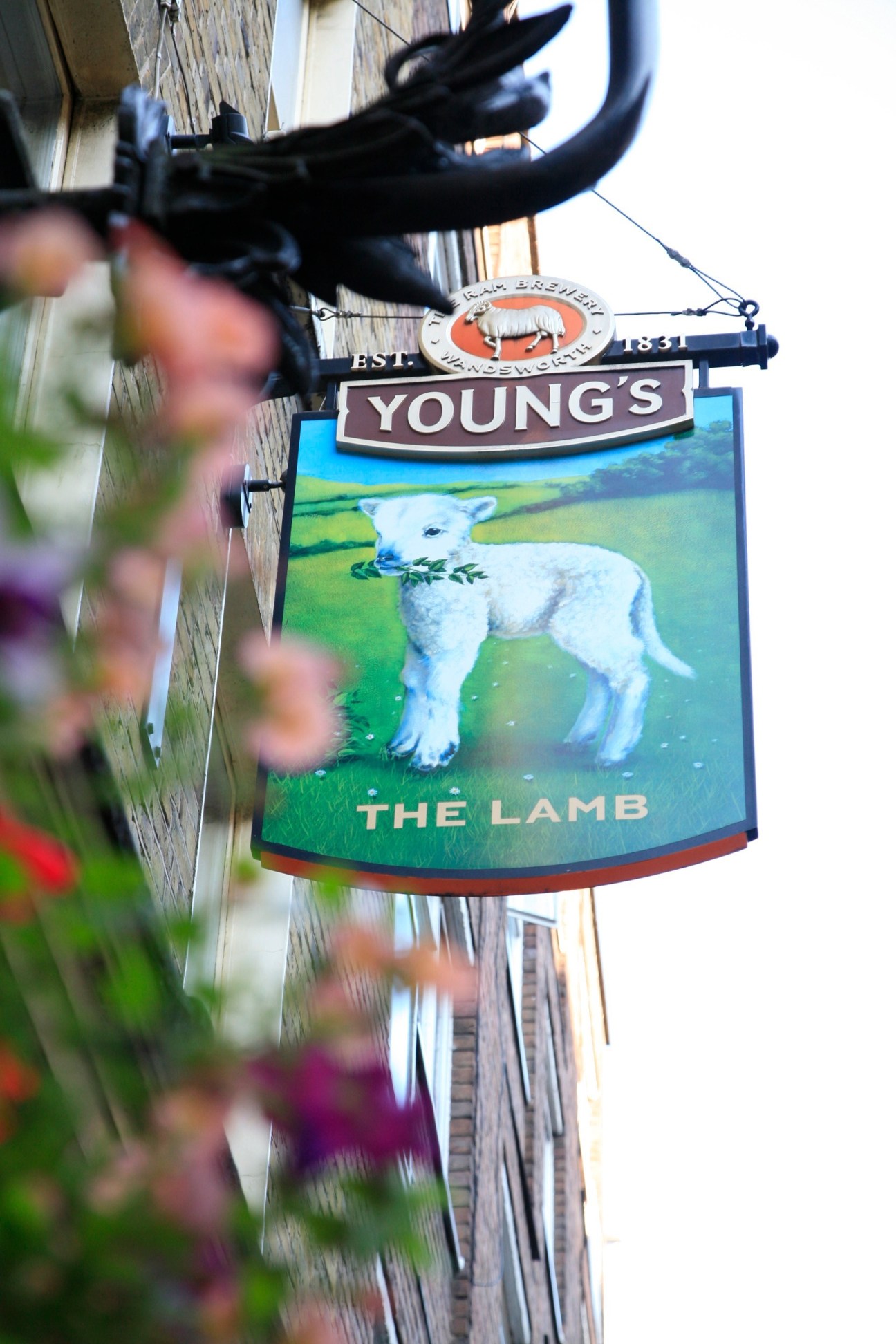 Young's pub sign for The Lamb featuring a white lamb with foliage, mounted on a brick building with flowers in foreground.