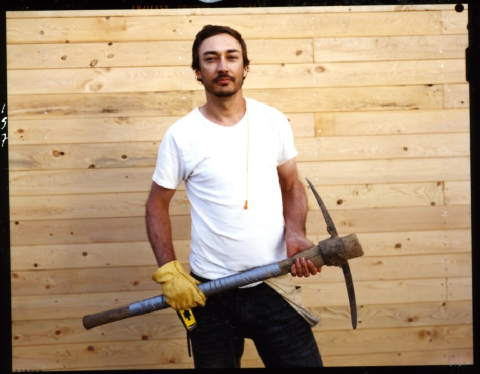Syracuse Masters student John Donalds holding a pickaxe against a wooden wall backdrop.