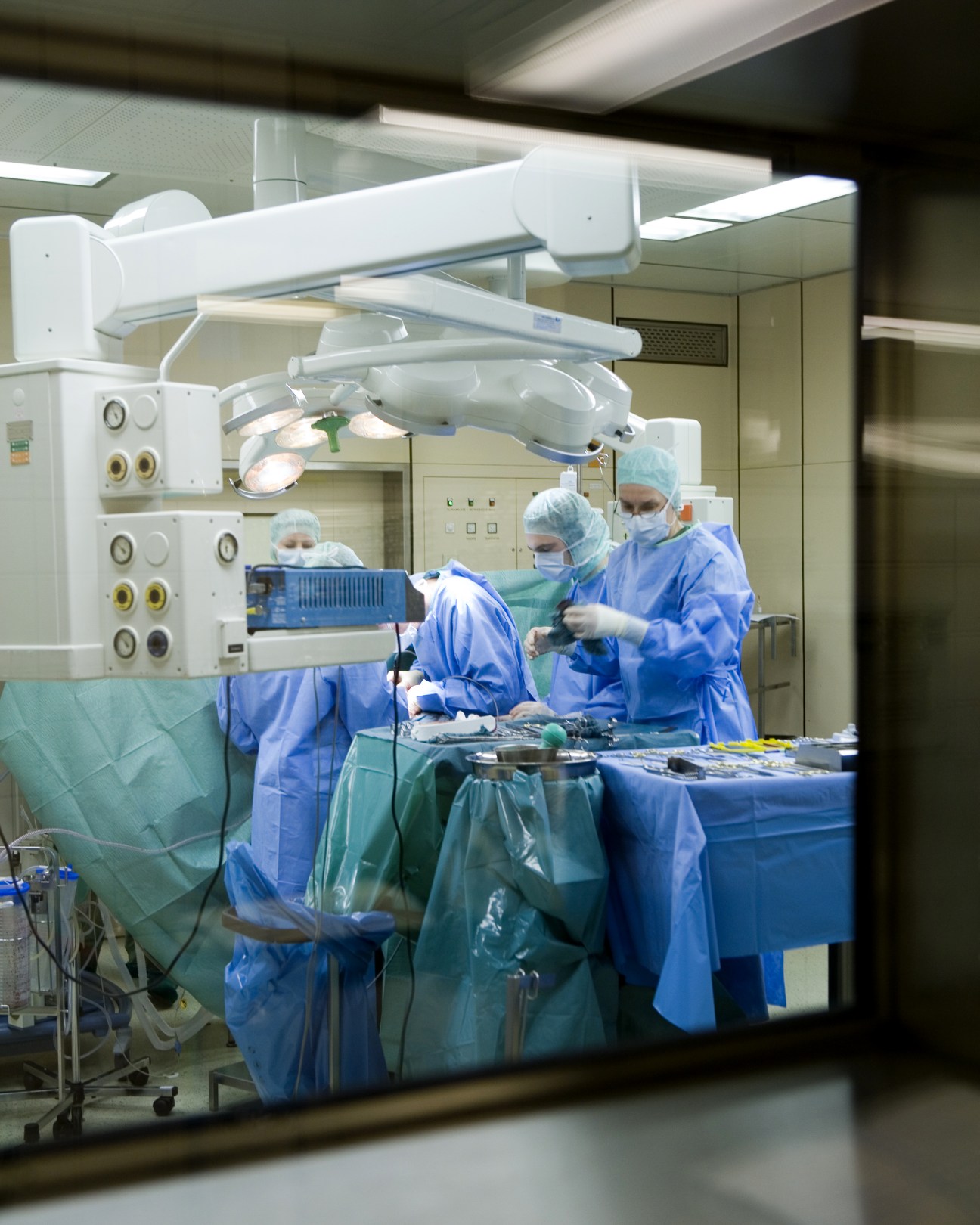 Surgeons in blue scrubs and masks perform surgery under bright operating lights in a modern hospital operating theatre.