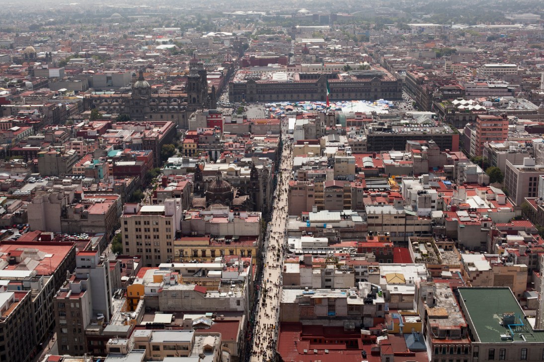Aerial view of Mexico City showing dense urban development with colonial architecture, red-tiled roofs, and a wide street lined with pedestrians.