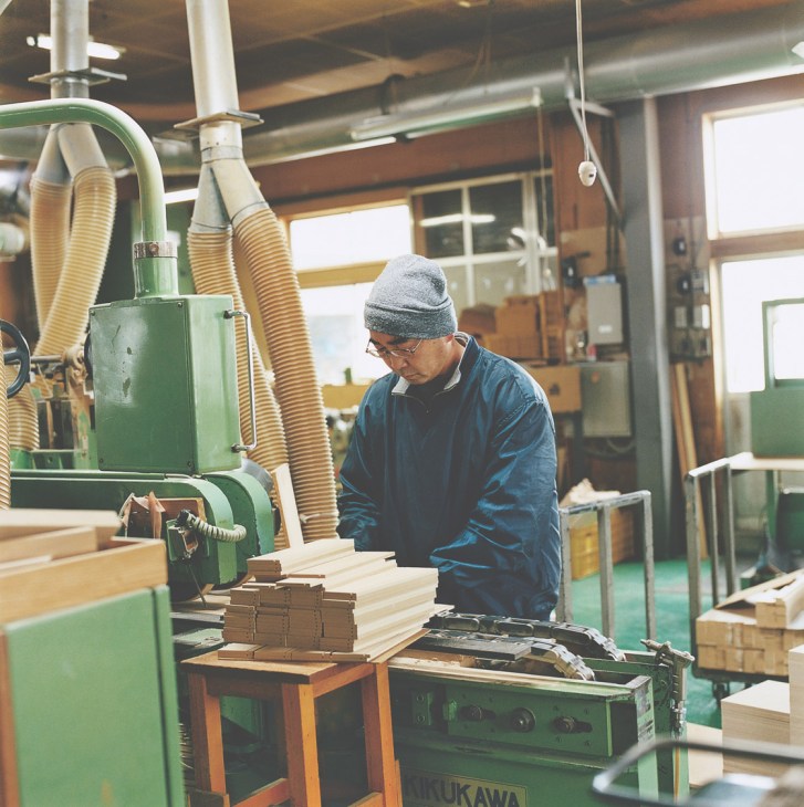 A woodworker trims wooden planks using a green Kuwahara machine in a bright workshop.