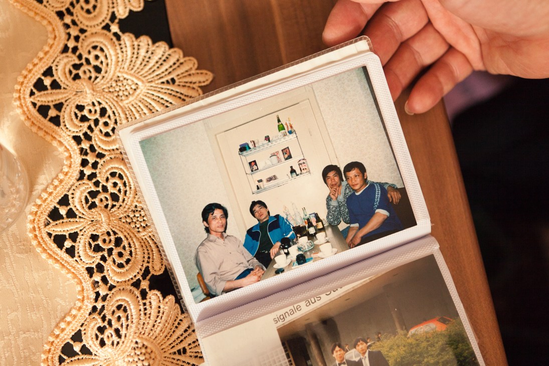 Hand holding a Polaroid photograph of four people gathered around a table with bottles, against a white shelf backdrop, with decorative lace fabric visible.