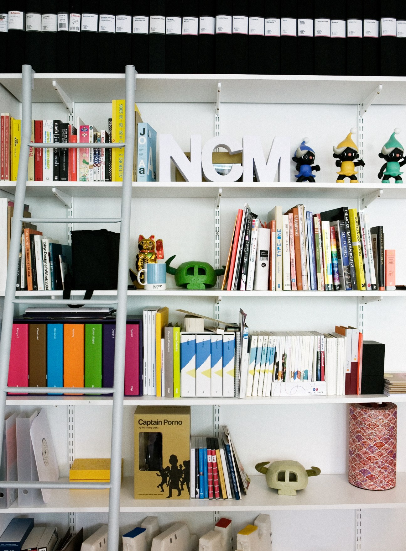 White shelving unit displaying colorful books, design objects, and toy figures in Next Century Modern office.