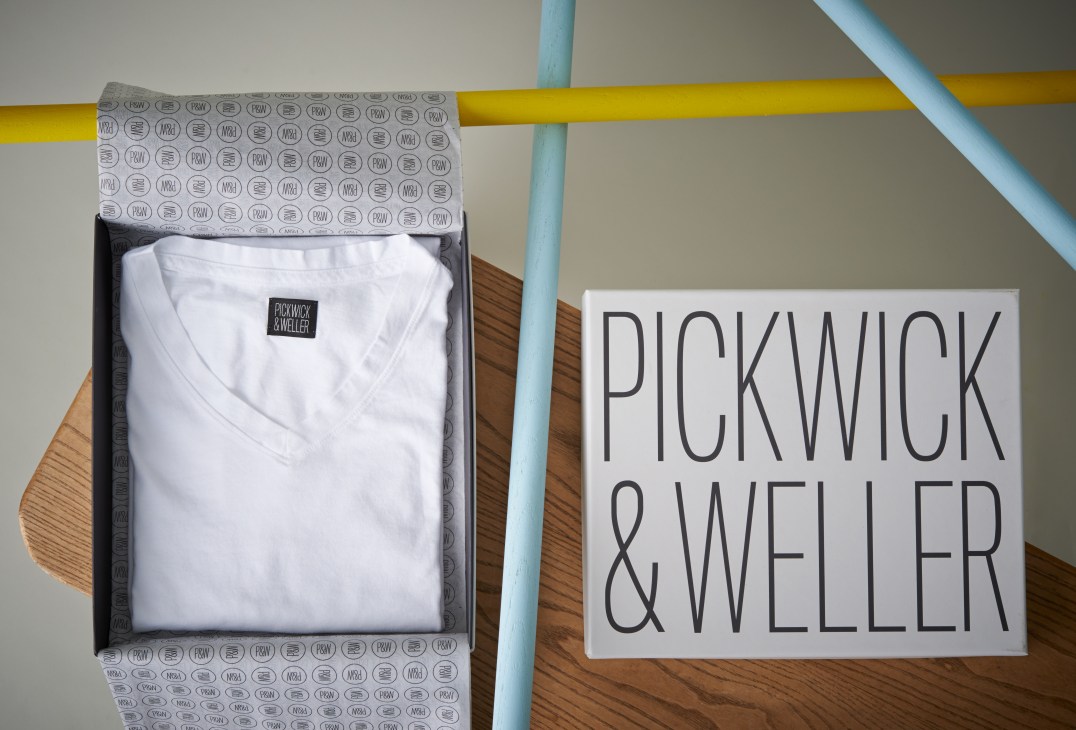 Pickwick & Weller branded t-shirt box with white folded t-shirt and signage on wooden surface with colored pipes.