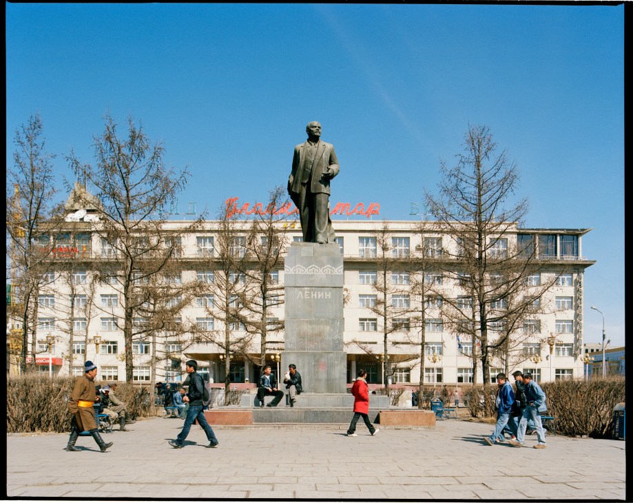 Statue of Lenin standing on a pedestal in a public square with Soviet-era apartment building behind it.