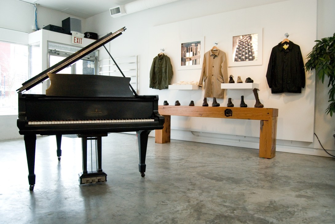 Lark shop interior with grand piano, wooden display table with boots, hanging jackets, and framed photographs on white walls.
